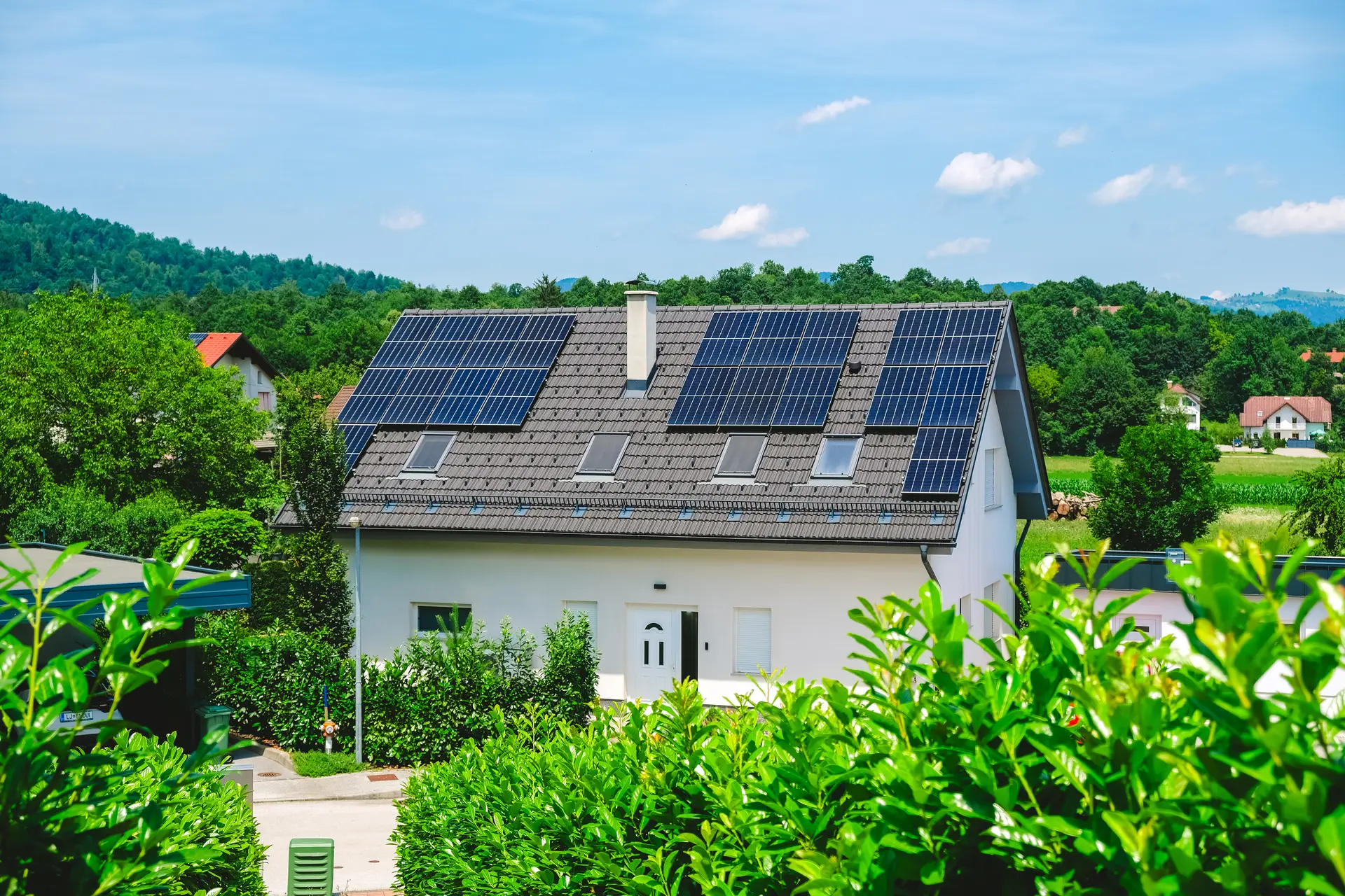 Weißes Einfamilienhaus mit Solarmodulen auf dem dunklen Ziegeldach, umgeben von grünen Hecken und bewaldeten Hügeln bei Sonnenschein